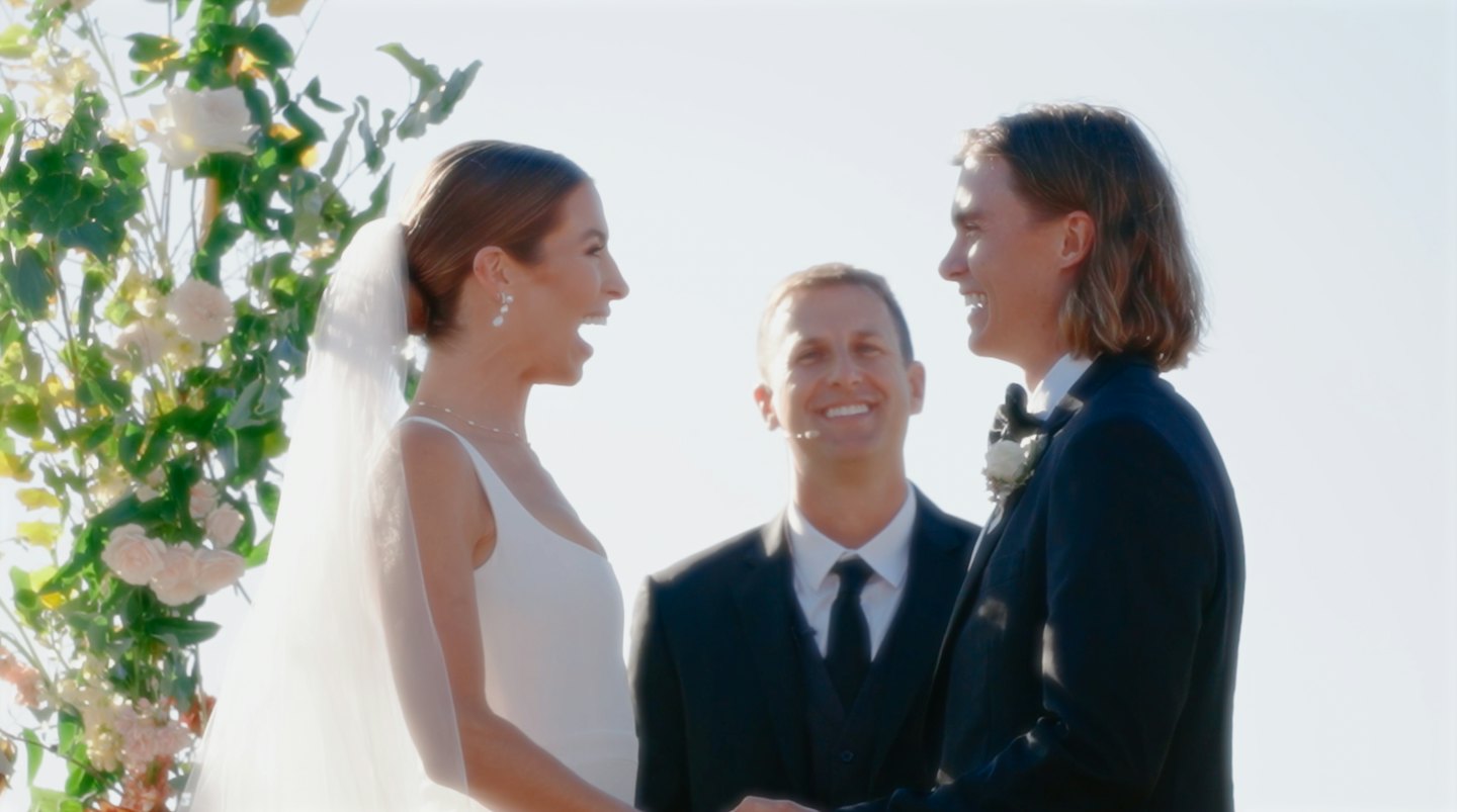 Bride and groom smile at each other under a floral arch.